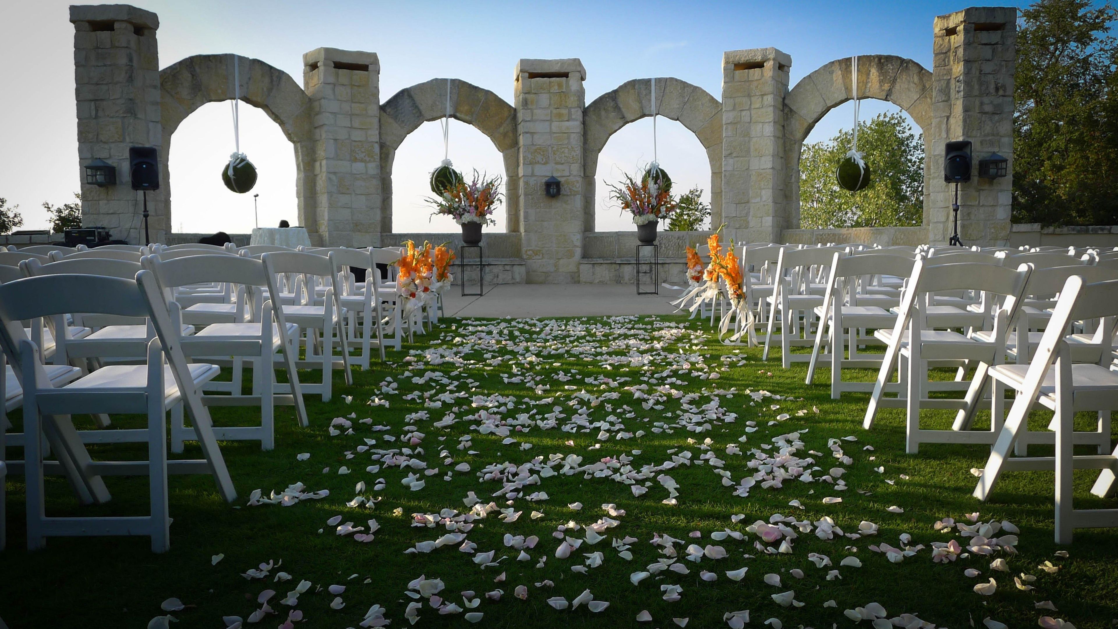 Un salón de bodas al aire libre con sillas blancas dispuestas en filas, arcos de piedra y pétalos de flores sobre el césped.