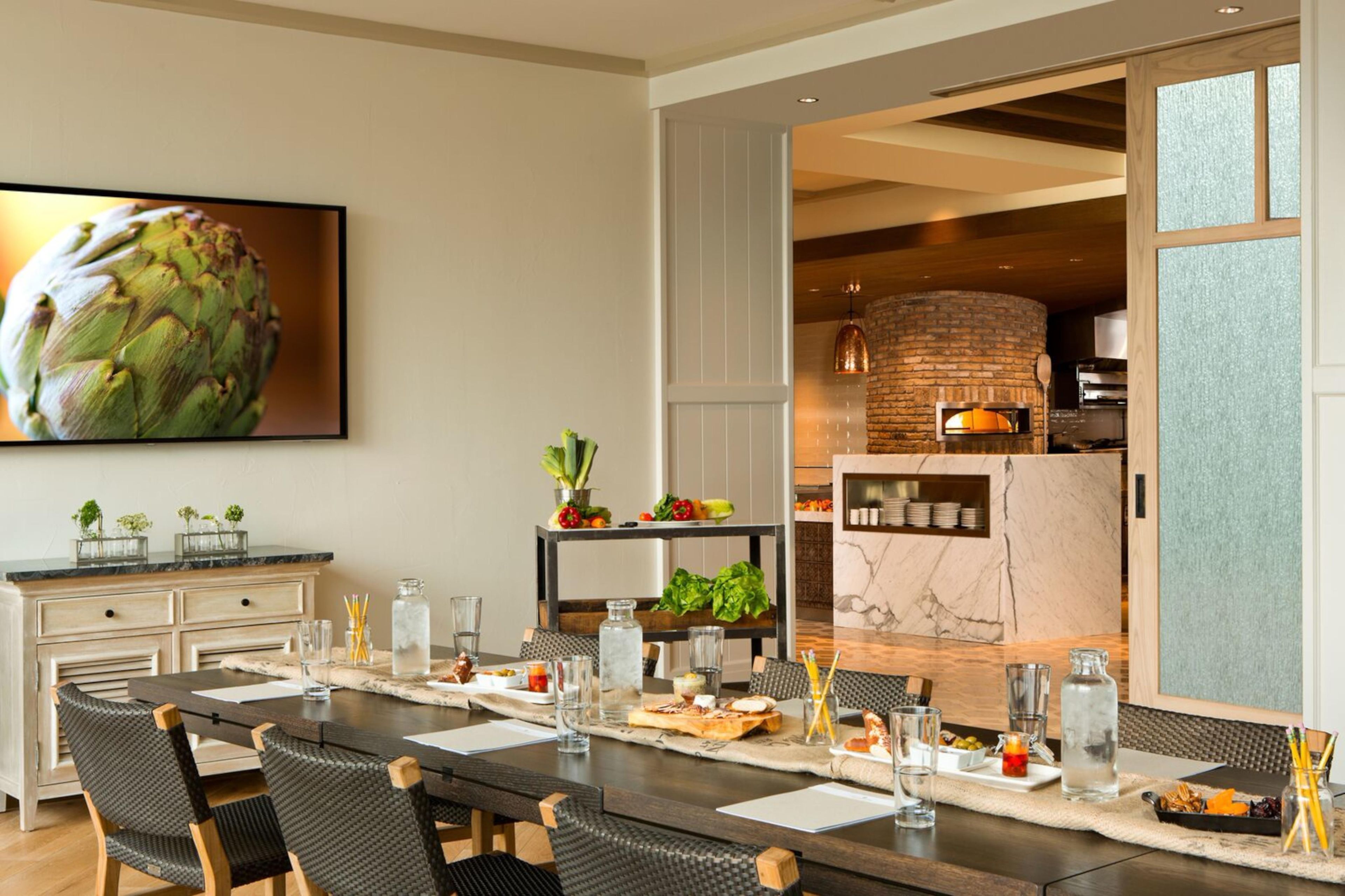 Un espace de repas moderne avec une longue table dressée pour un repas, une cuisine dotée d’un four en briques et une grande œuvre d’art encadrée au mur.