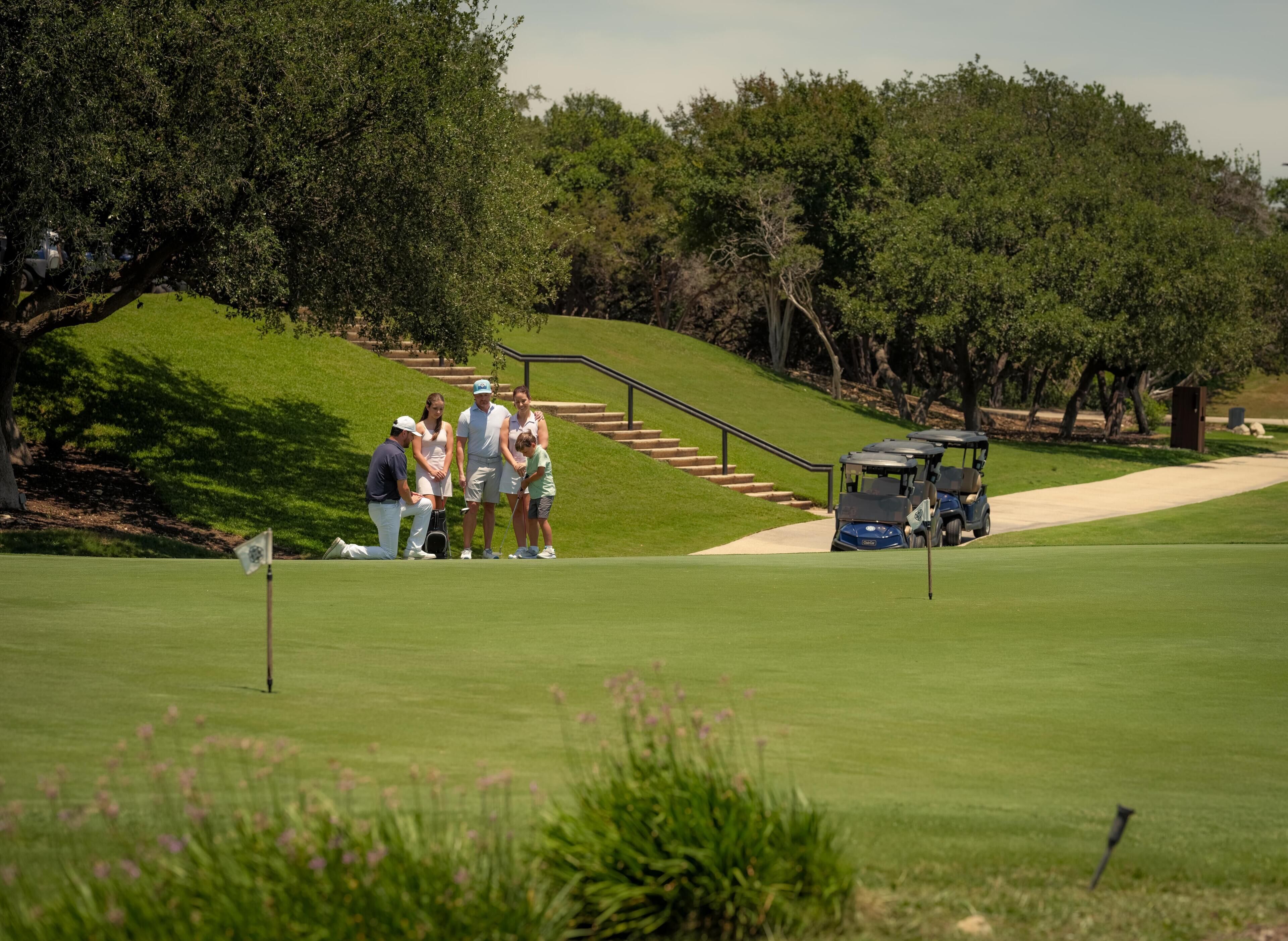 Ein Golfplatz mit einer Gruppe von Personen in der Nähe eines Flaggestangs, einem Golfwagen und einer Treppe, die einen Hügel hinaufführt.