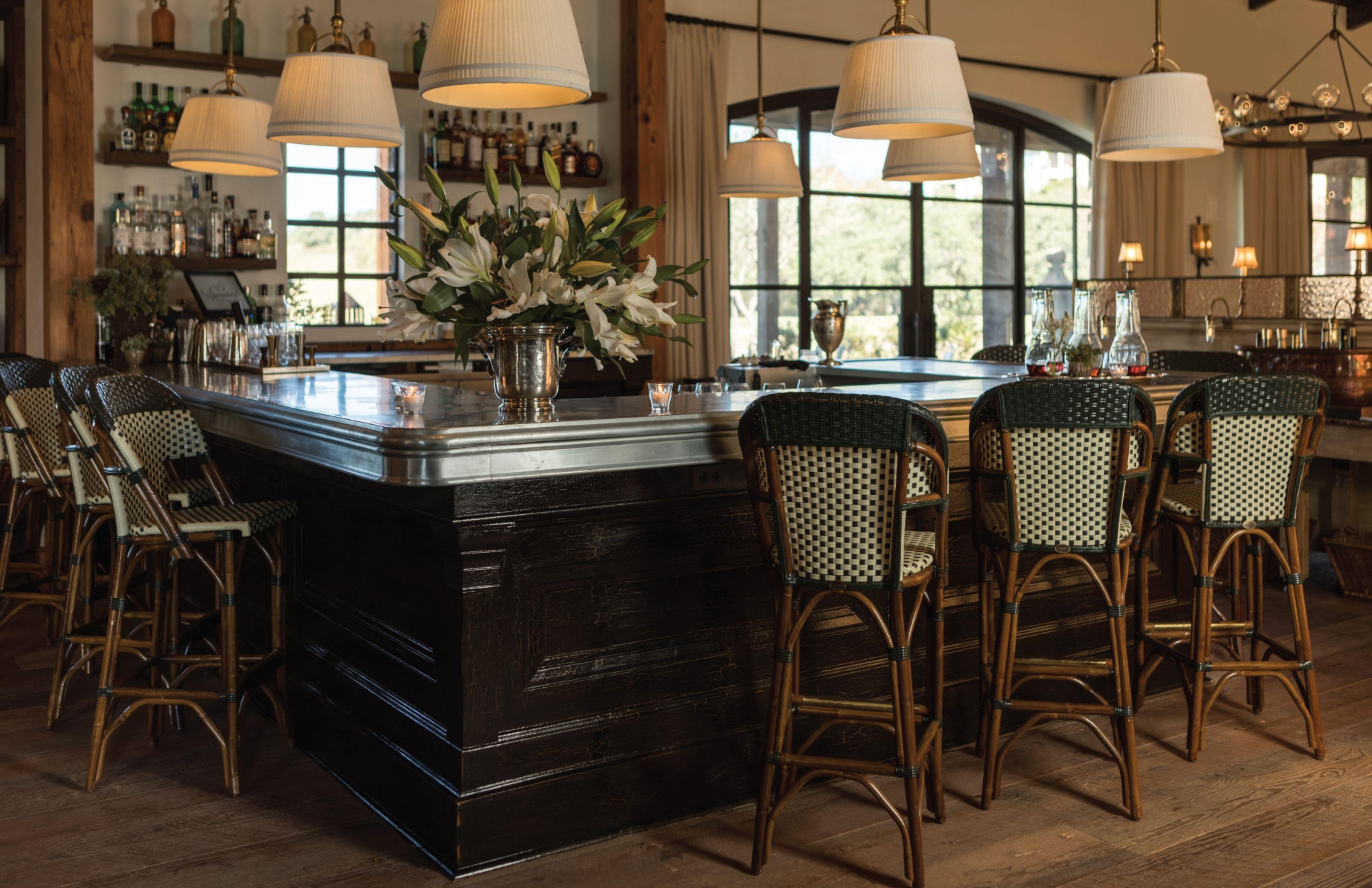 A bar area with a central island, bar stools, hanging lamps, and a view of the outdoors through large windows.