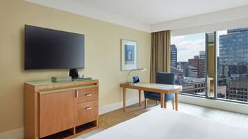 A hotel room with a large TV, a desk with a lamp, a chair, and a view of the cityscape.