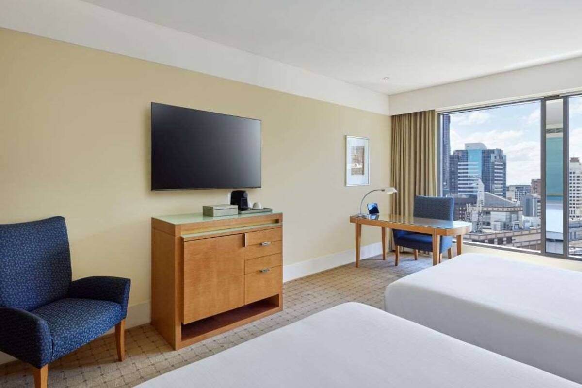 Ein Hotelzimmer mit Bett, Schreibtisch, Stuhl, Fernseher und einem großen Fenster mit Blick auf die Stadt.