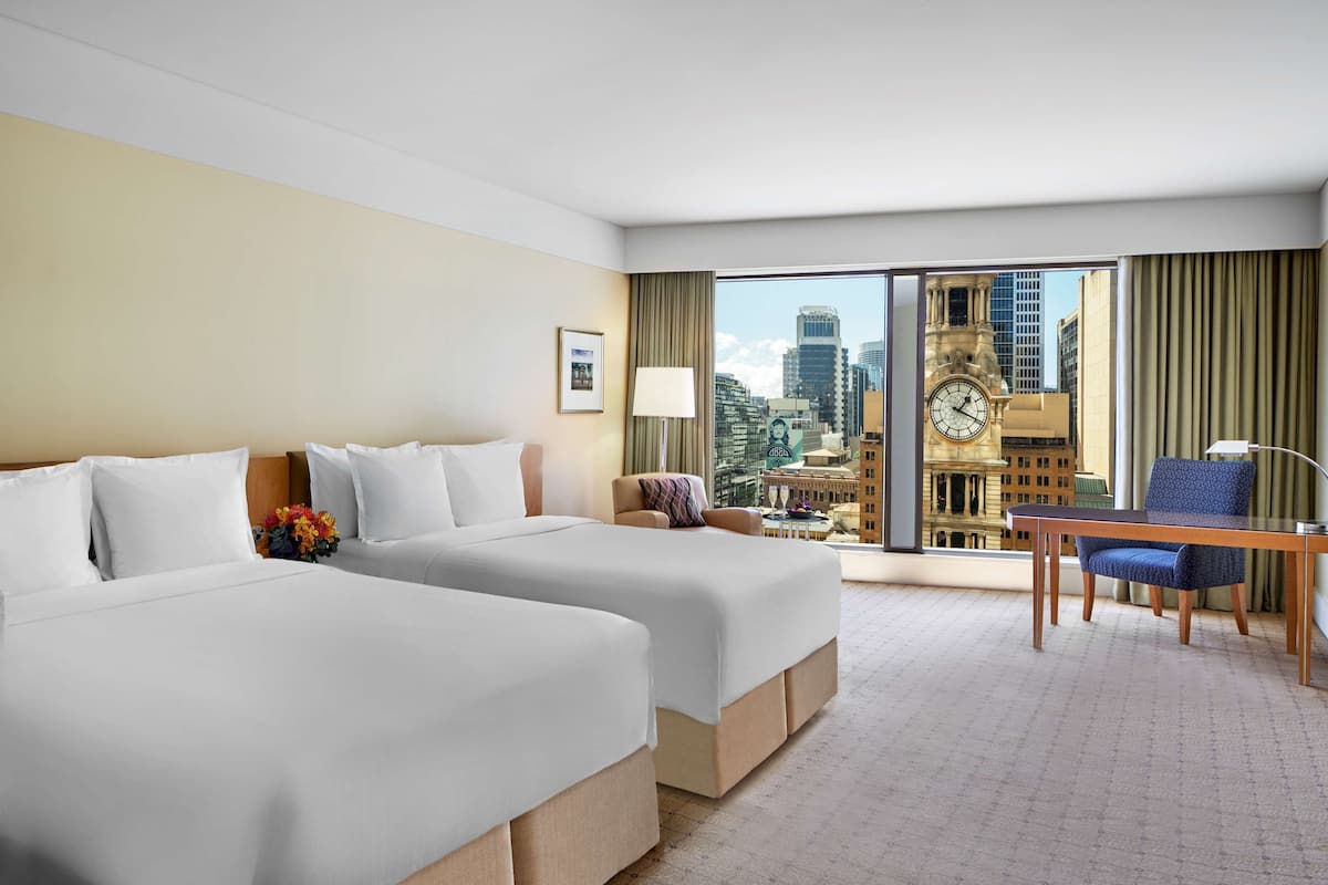 Ein Hotelzimmer mit zwei Betten, einem Schreibtisch, einem Stuhl und Blick auf eine Stadtlandschaft mit einem Glockenturm.