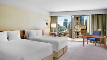 Une chambre d’hôtel avec deux lits, un bureau, une chaise et une vue sur le paysage urbain, notamment sur un beffroi.