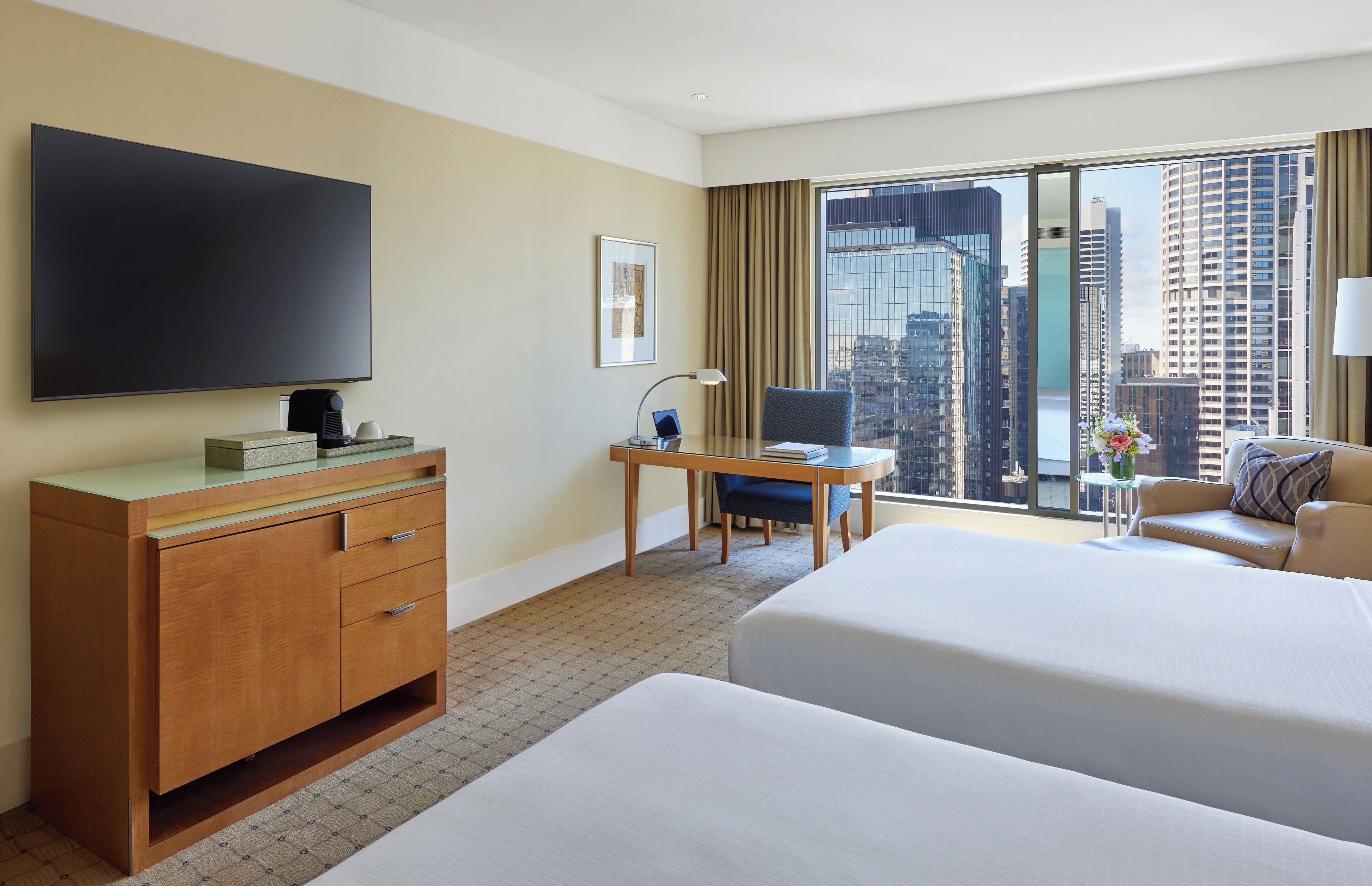 Habitación de hotel con dos camas, un escritorio, un televisor, una ventana con vista a la ciudad y una mesita de noche con lámpara.