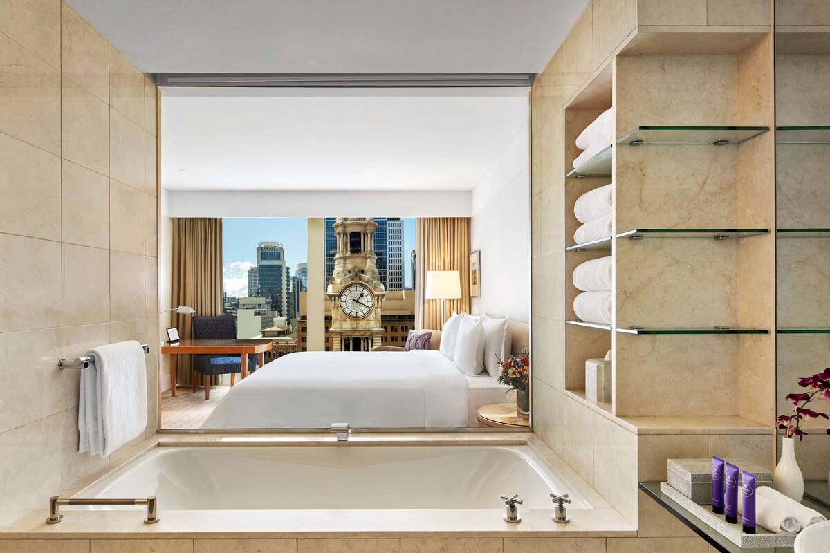 Ein Hotelzimmer mit einer großen Badewanne, einem Bett, einem Schreibtisch mit Stuhl, einer Uhr und Blick auf die Stadt.