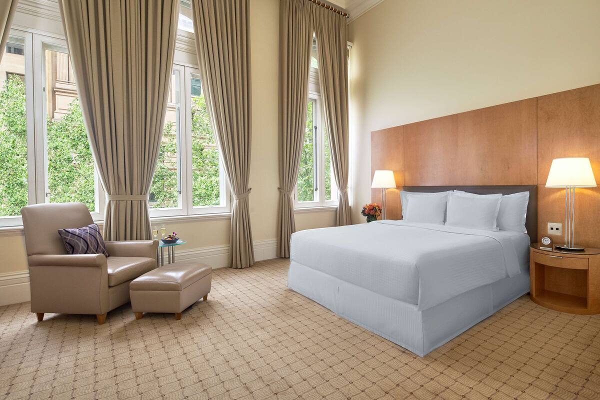 Ein Hotelzimmer mit einem großen Bett, einem Sessel, einem Hocker, einem Nachttisch und einer Lampe.
