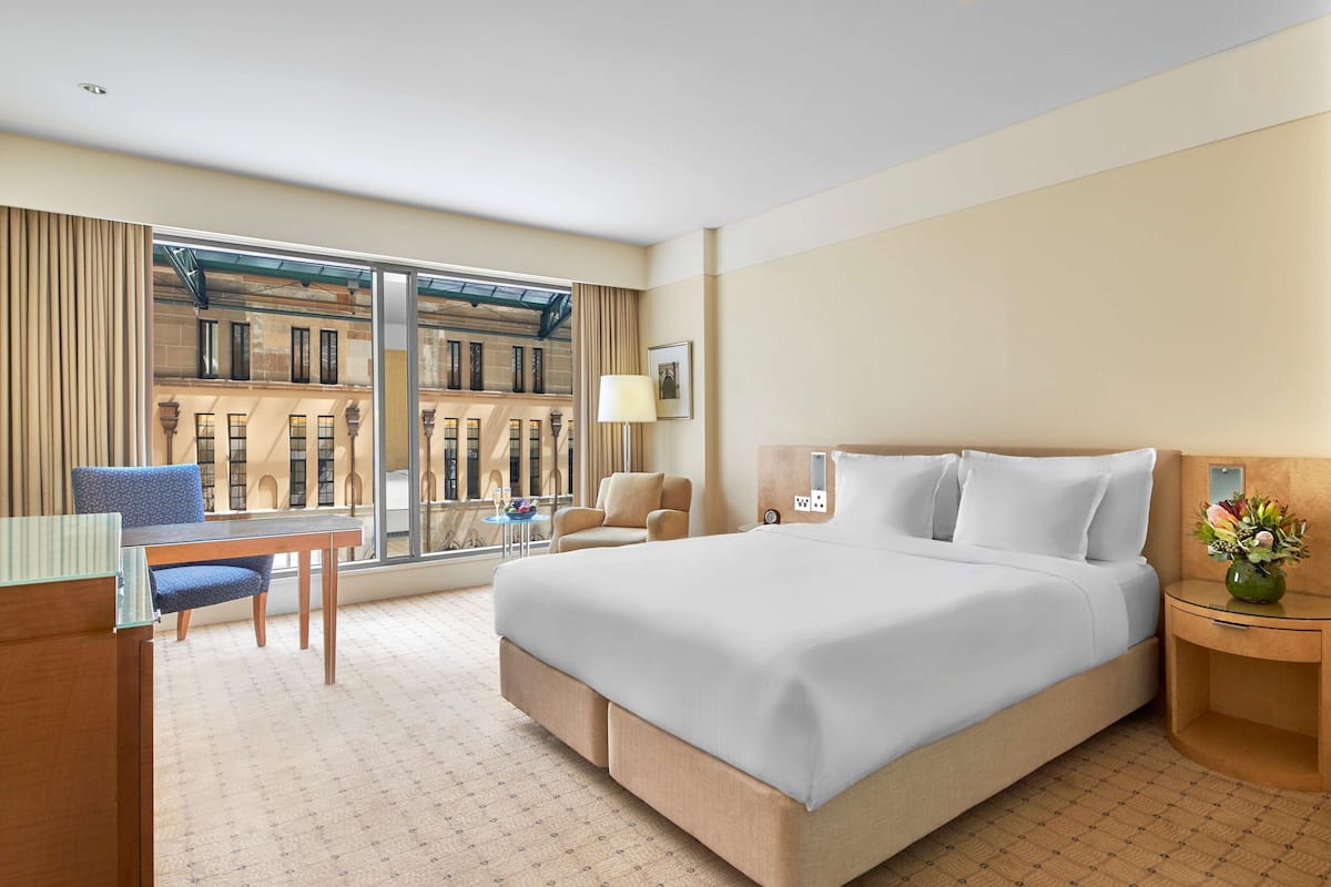 Ein Hotelzimmer mit einem großen Bett, einem Schreibtisch, einem Sessel, einem kleinen Tisch und Blick auf ein Gebäude durch große Fenster.
