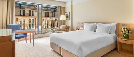 Habitación de hotel con una cama grande, un escritorio, una silla, una mesita y vistas a un edificio a través de amplias ventanas.