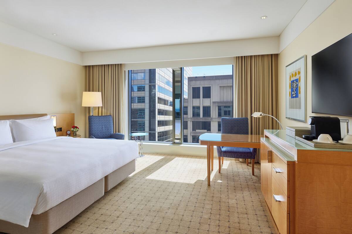 Ein Hotelzimmer mit einem großen Bett, einem Schreibtisch mit Stuhl, einem Fernseher und Blick auf die Stadt durch das Fenster.