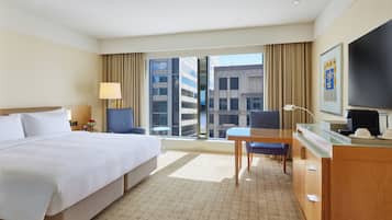 A hotel room with a large bed, a desk with a chair, a television, and a view of the city through the window.