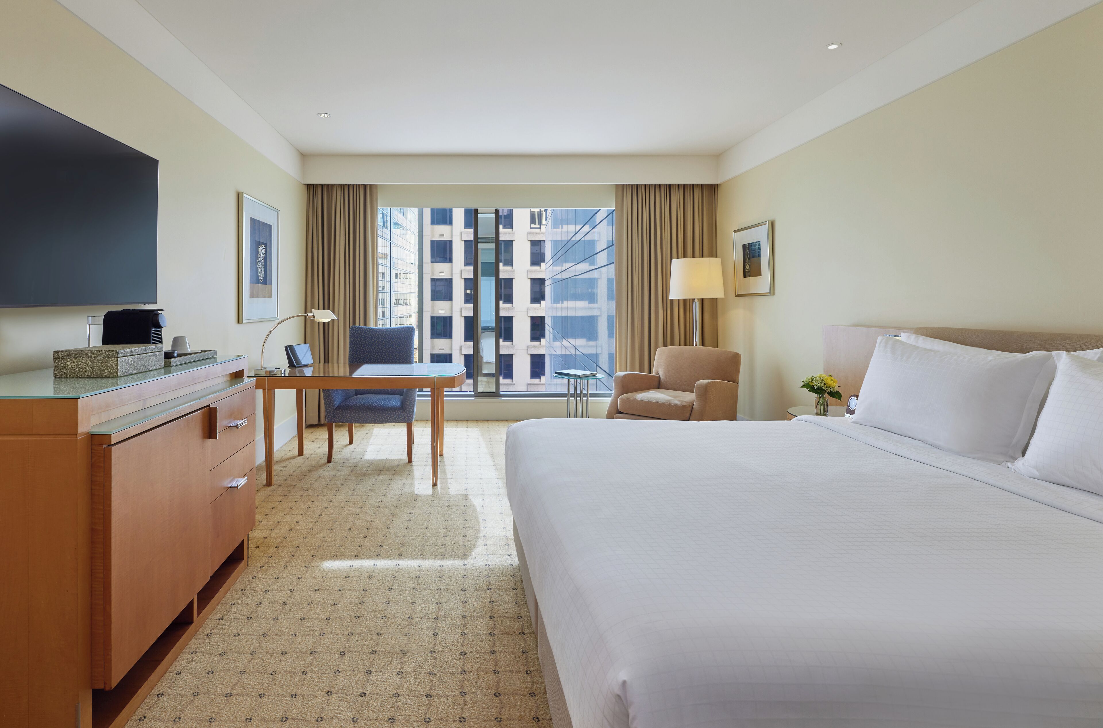 A hotel room with a large bed, a desk with a chair, a TV, and a window with a view of buildings.