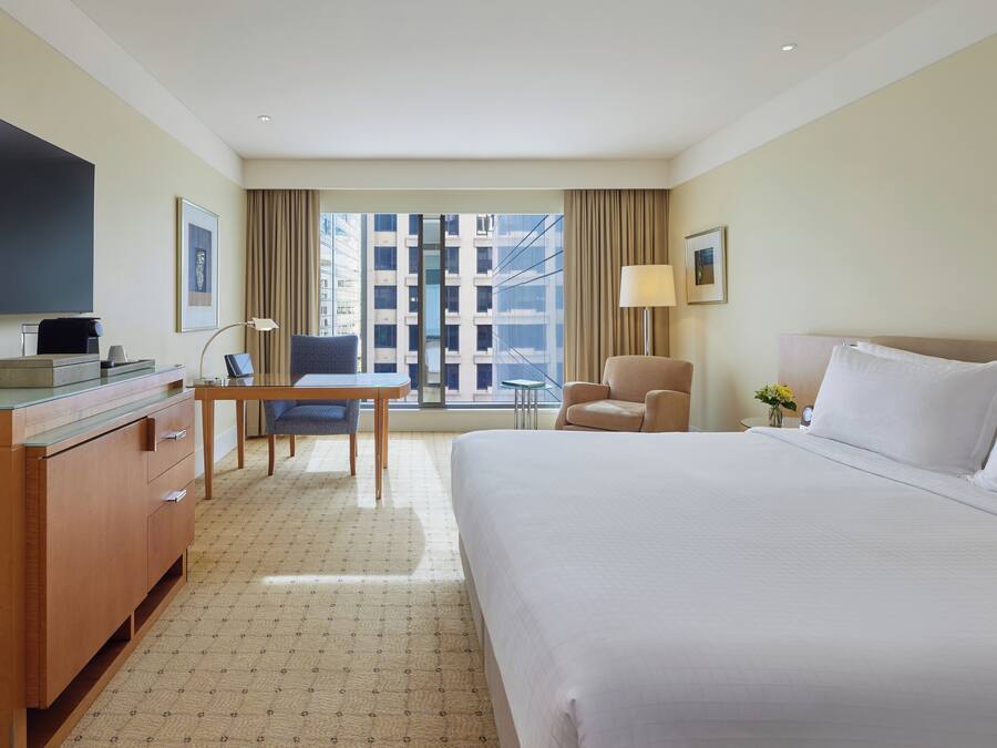 A hotel room with a large bed, a desk with a chair, a TV, and a window with a view of buildings.
