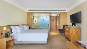 A hotel room with a large bed, a desk with a flat-screen TV, a wooden dresser, and a bathroom visible through an open door.
