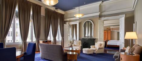 Una habitación de hotel con alfombra azul, chimenea, cama, zona de estar con sillas y ventanas grandes con cortinas.