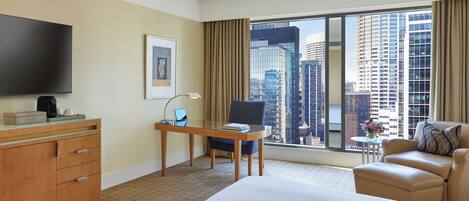 Habitación de hotel con cama, escritorio con computadora portátil, silla, televisor y vista a edificios de la ciudad.