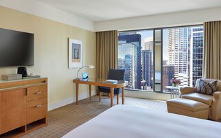 Habitación de hotel con cama, escritorio con computadora portátil, silla, televisor y vista a edificios de la ciudad.