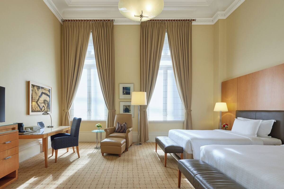 Ein Hotelzimmer mit zwei Betten, einem Schreibtisch, einem Sessel, einer Lampe und einem Fernseher.