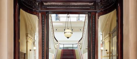 A grand staircase with a red carpet leading to a high-ceilinged area with ornate woodwork and a hanging light fixture.
