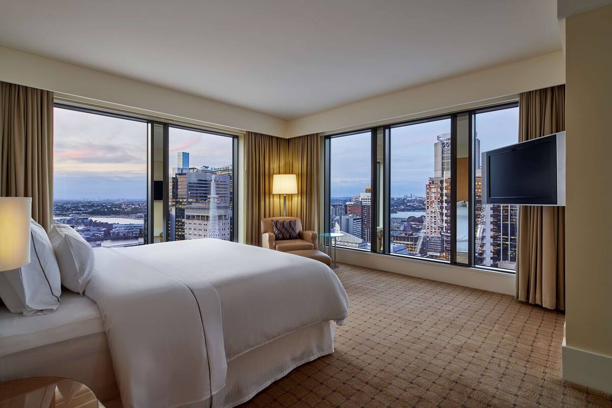 Ein Hotelzimmer mit einem großen Bett, einem Sessel, einem Fernseher und Blick auf die Skyline durch große Fenster.