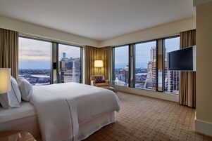 Habitación de hotel con una cama grande, una silla, un televisor y vistas a la ciudad a través de amplios ventanales.