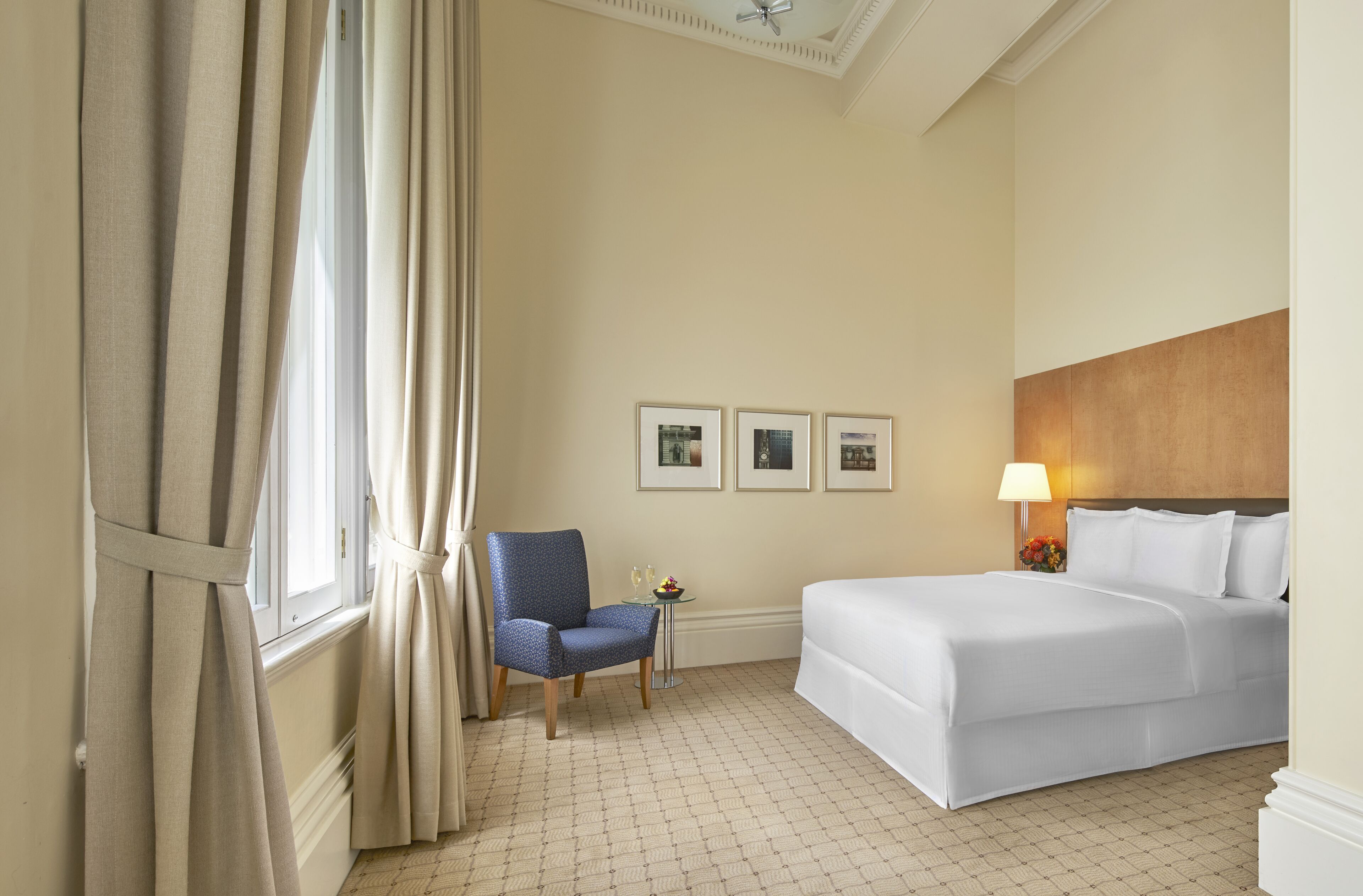 Une chambre d’hôtel avec un lit, une chaise, une petite table avec une lampe et trois tableaux encadrés accrochés au mur.