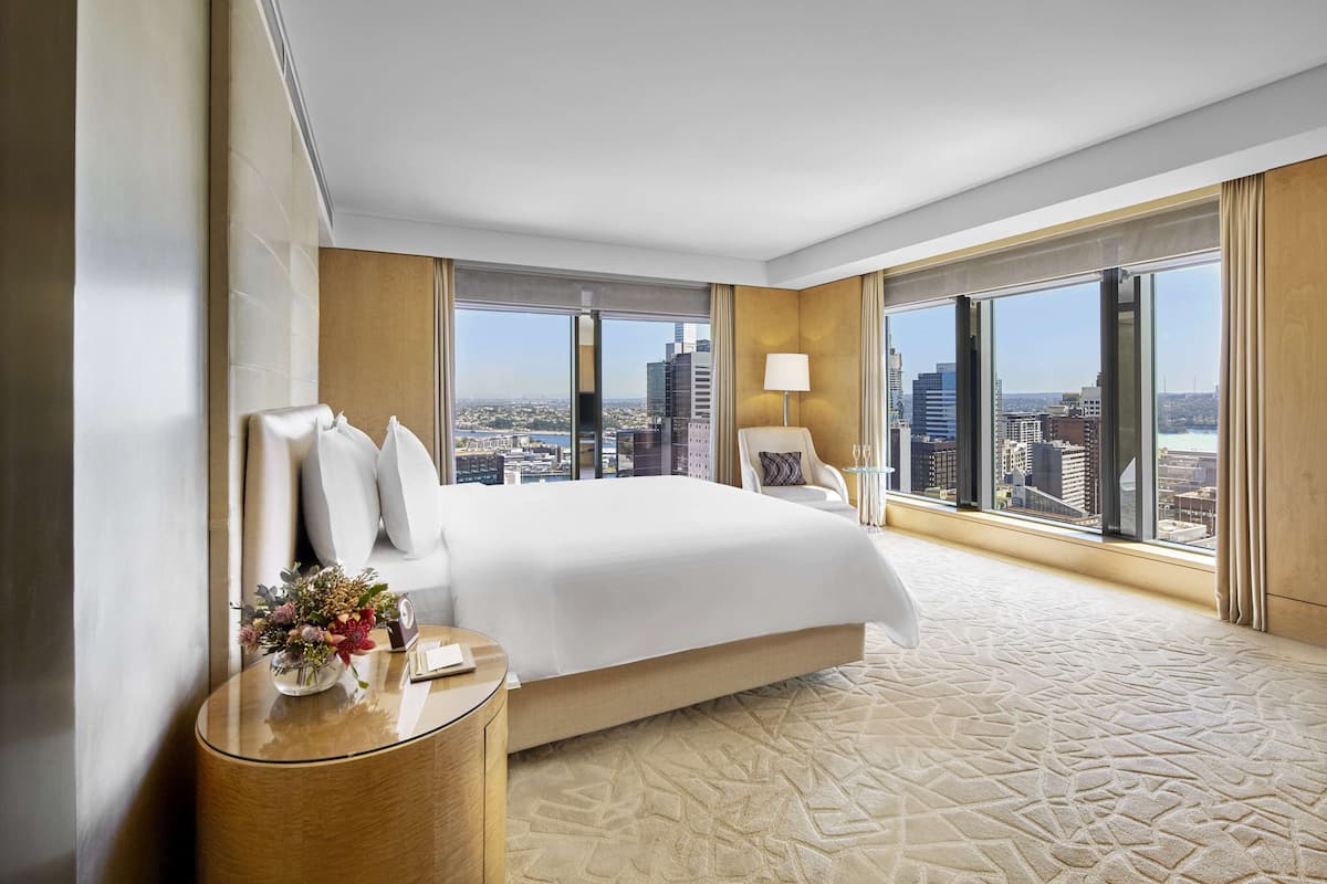 Ein modernes Hotelzimmer mit einem großen Bett, einem Nachttisch mit Blumen in einer Vase, einer Lampe und einem Blick auf die Stadt durch große Fenster.
