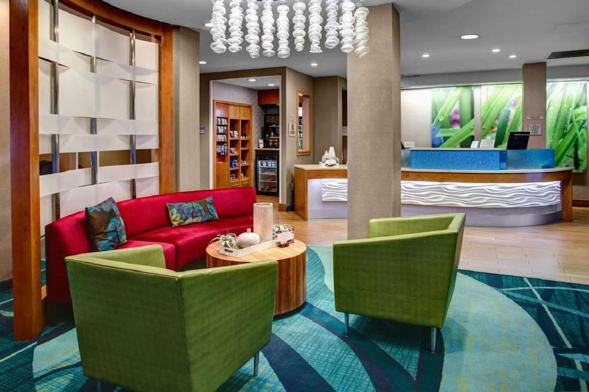 Een hotel lobby met een receptiebalie, een rode bank, groene fauteuils, een bijzettafeltje en een kamerbreed tapijt.