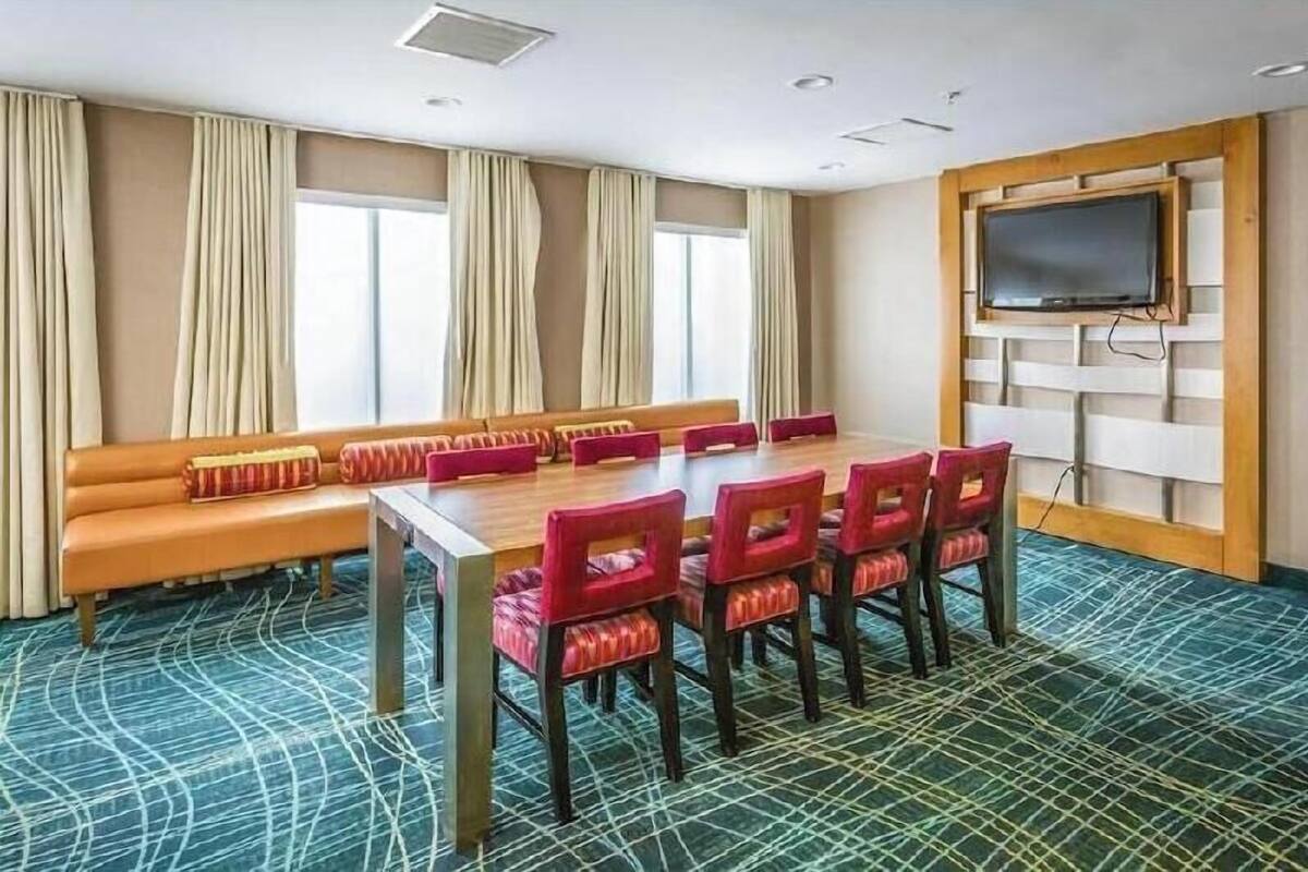 A conference room with a long table, red chairs, a sofa, beige curtains, a television, and patterned carpet.