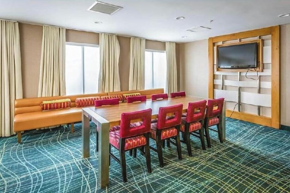 A conference room with a long table, red chairs, a sofa, beige curtains, a television, and patterned carpet.