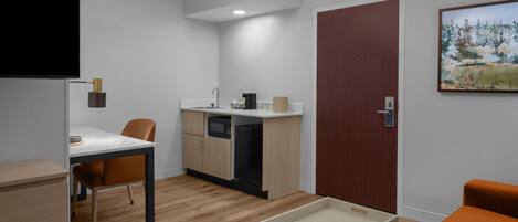 A modern room with a wooden floor, a dining table with chairs, a kitchenette, a wooden door, and a painting on the wall.