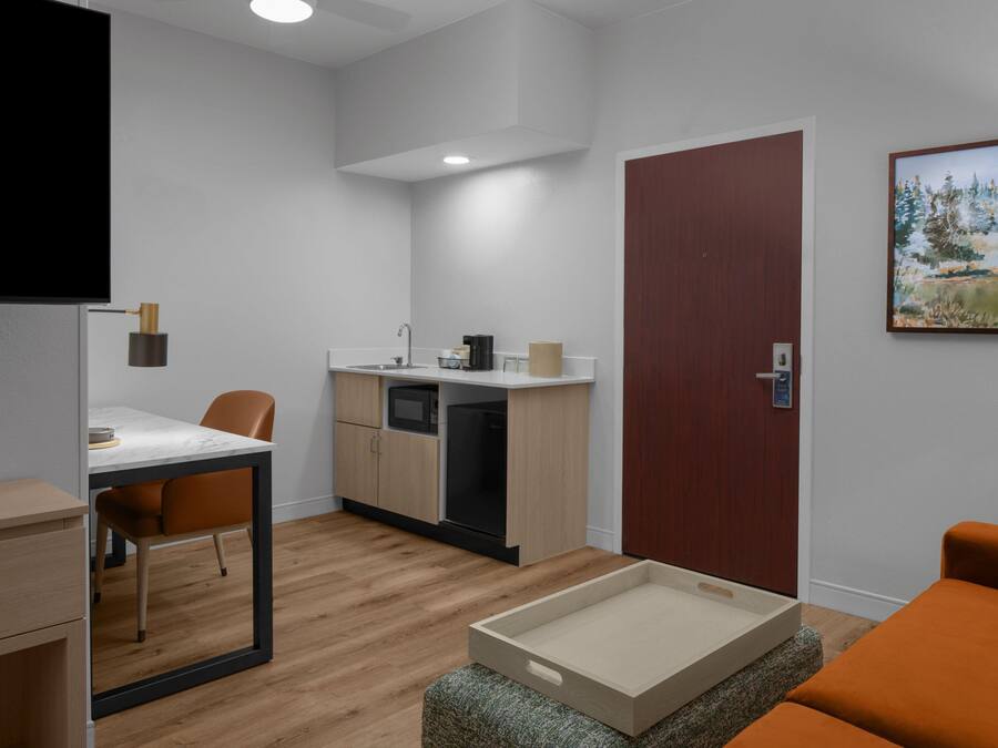 A modern room with a wooden floor, a dining table with chairs, a kitchenette, a wooden door, and a painting on the wall.