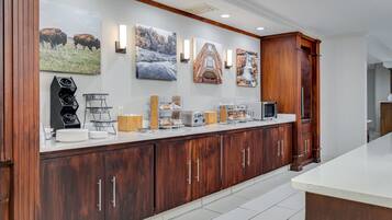 A modern kitchen with wooden cabinets, a white countertop, and various appliances. There are wall-mounted light fixtures and artwork depicting nature scenes.