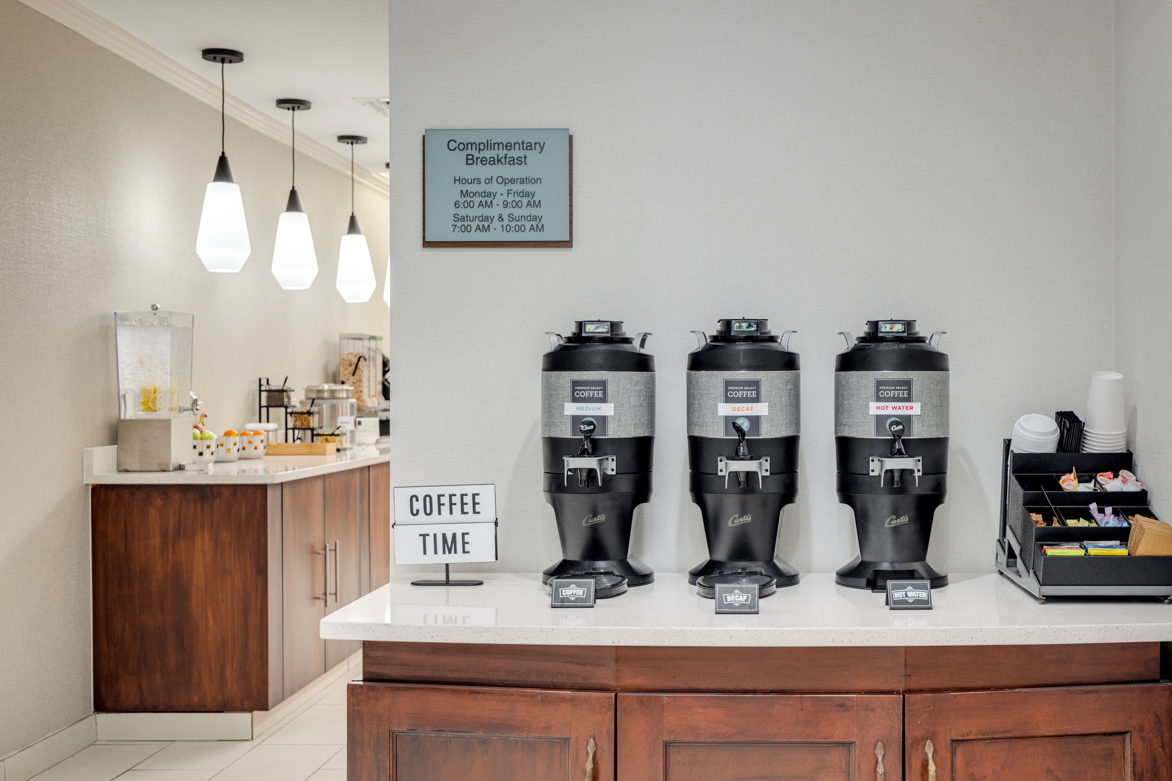 Una estación de café con tres dispensadores, un letrero que indica desayuno de cortesía y un letrero con el horario del café.