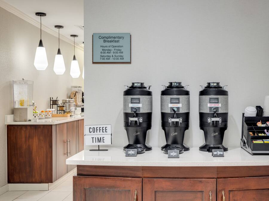 Uma estação de café com três dispensadores, um aviso de café da manhã cortesia e um aviso com o horário do café.
