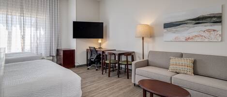 Habitación de hotel con cama, sofá, escritorio con sillas, televisor y un cuadro en la pared.
