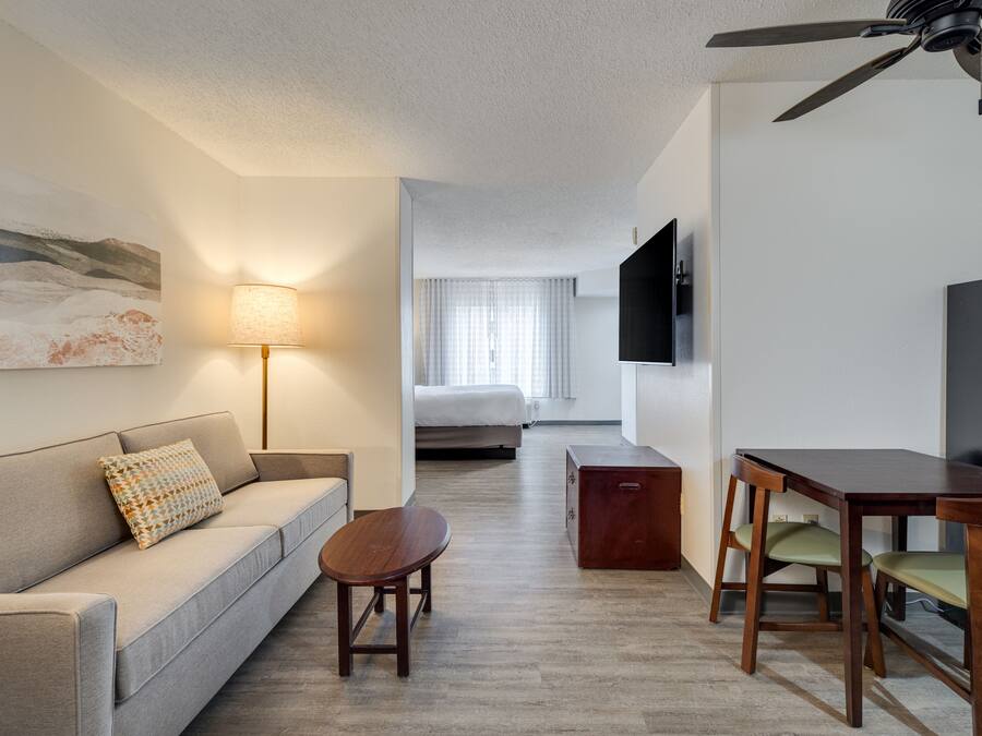 A hotel room with a sofa, a small wooden table, a desk with chairs, a flat-screen TV, a ceiling fan, and a bed with a window.