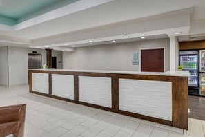 A reception area with a wooden counter, white decorative panels, a refrigerator with beverages, and a door in the background.