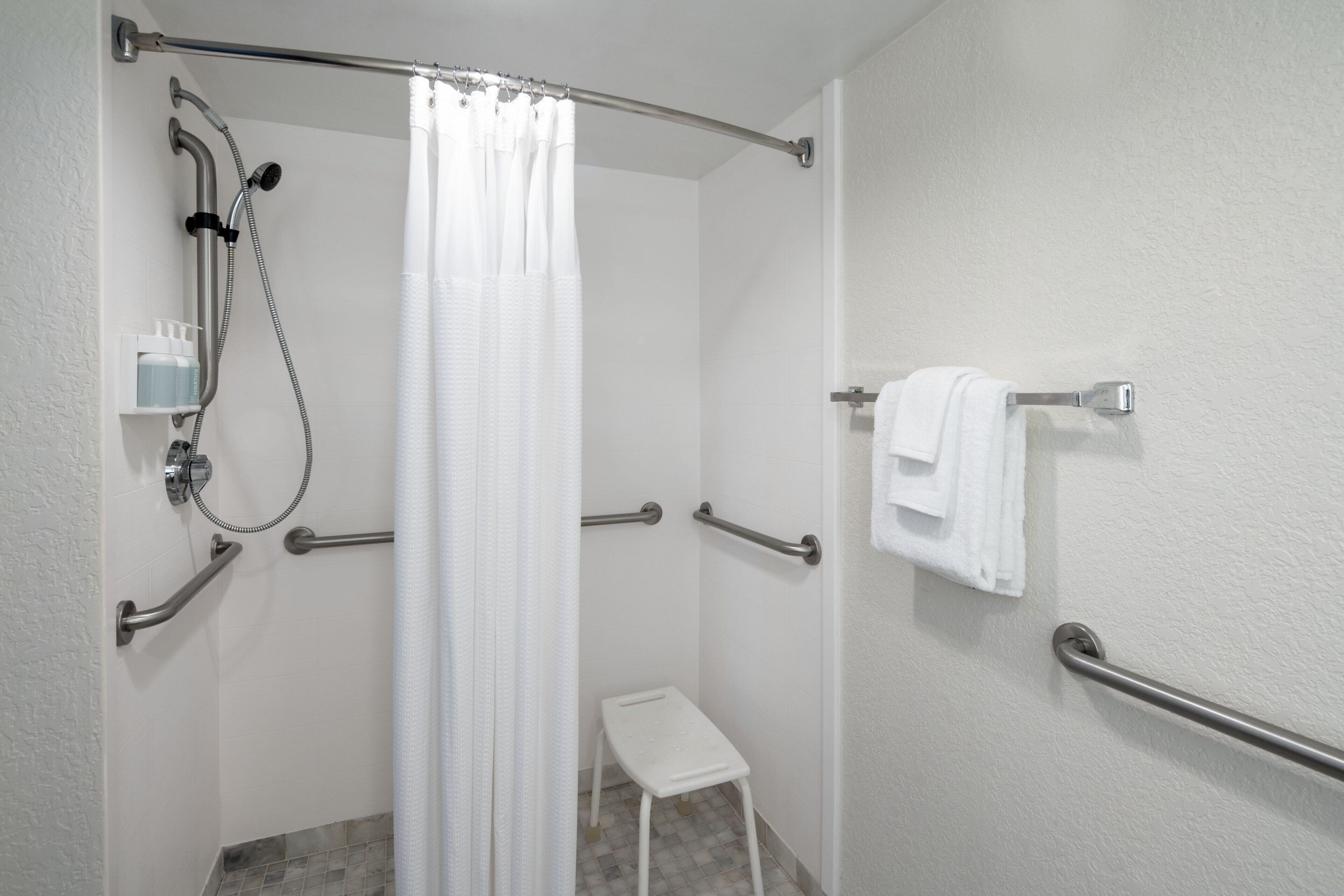 A bathroom with a shower, grab bars, a white towel rack, and a small step stool.
