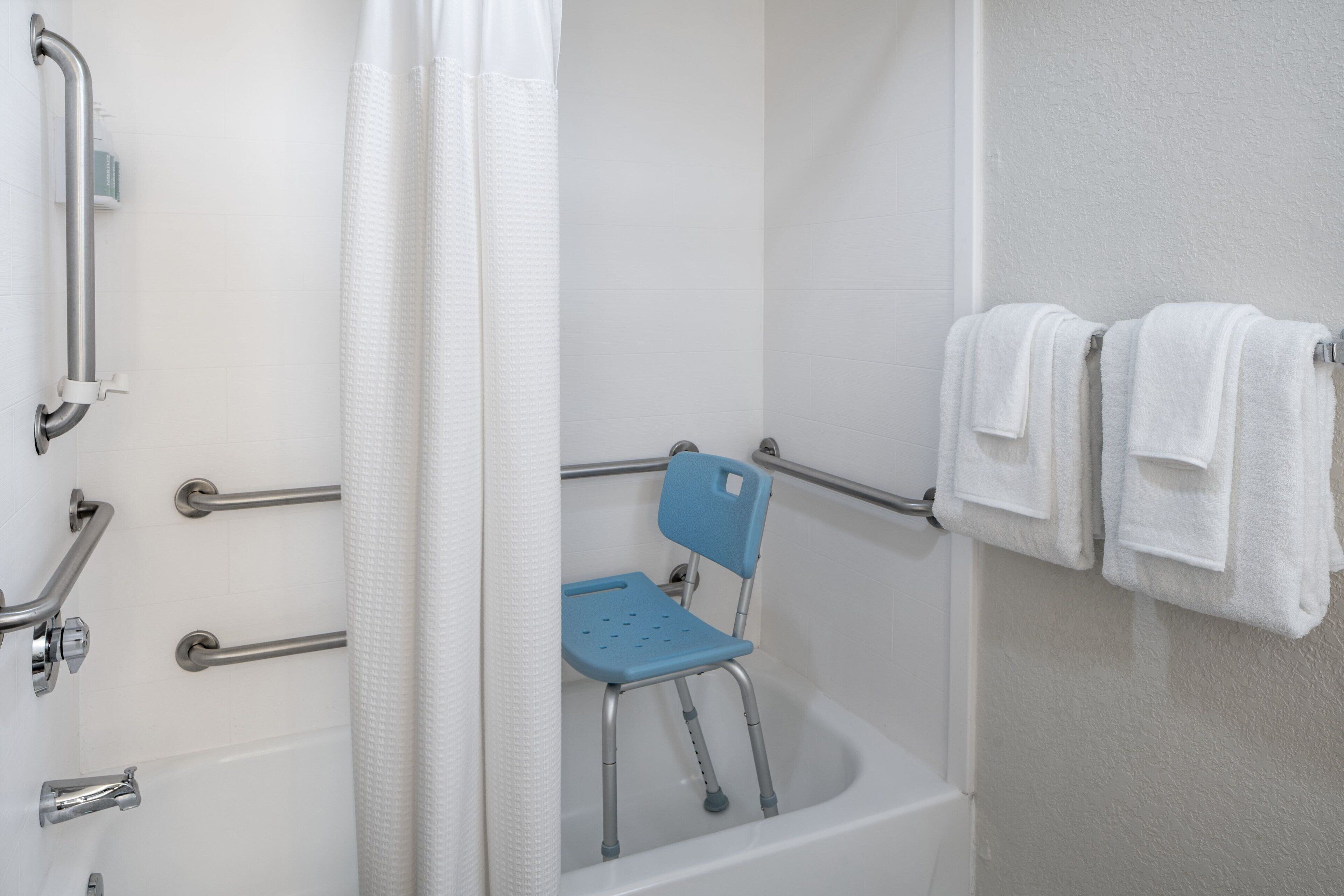 Un baño con bañera, barras de apoyo, un asiento para la ducha y toallas en un perchero.
