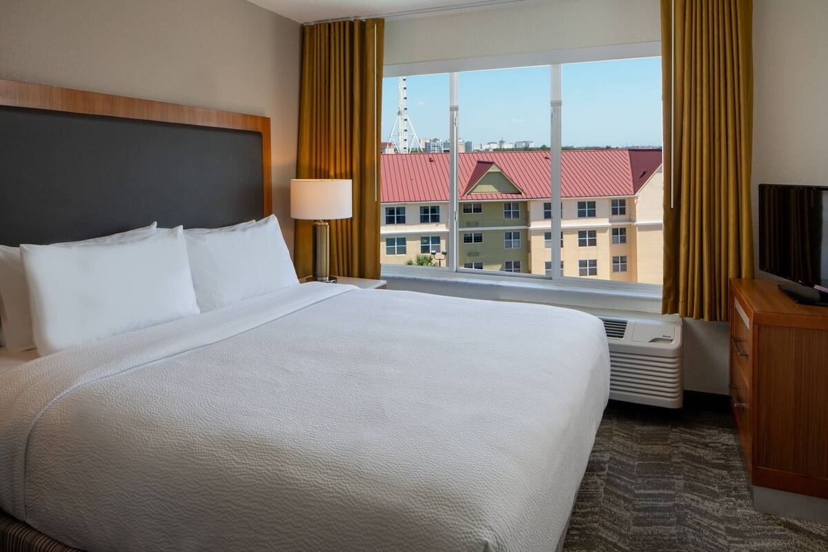 A hotel room with a large bed, a TV, a window with curtains, and a view of buildings outside.