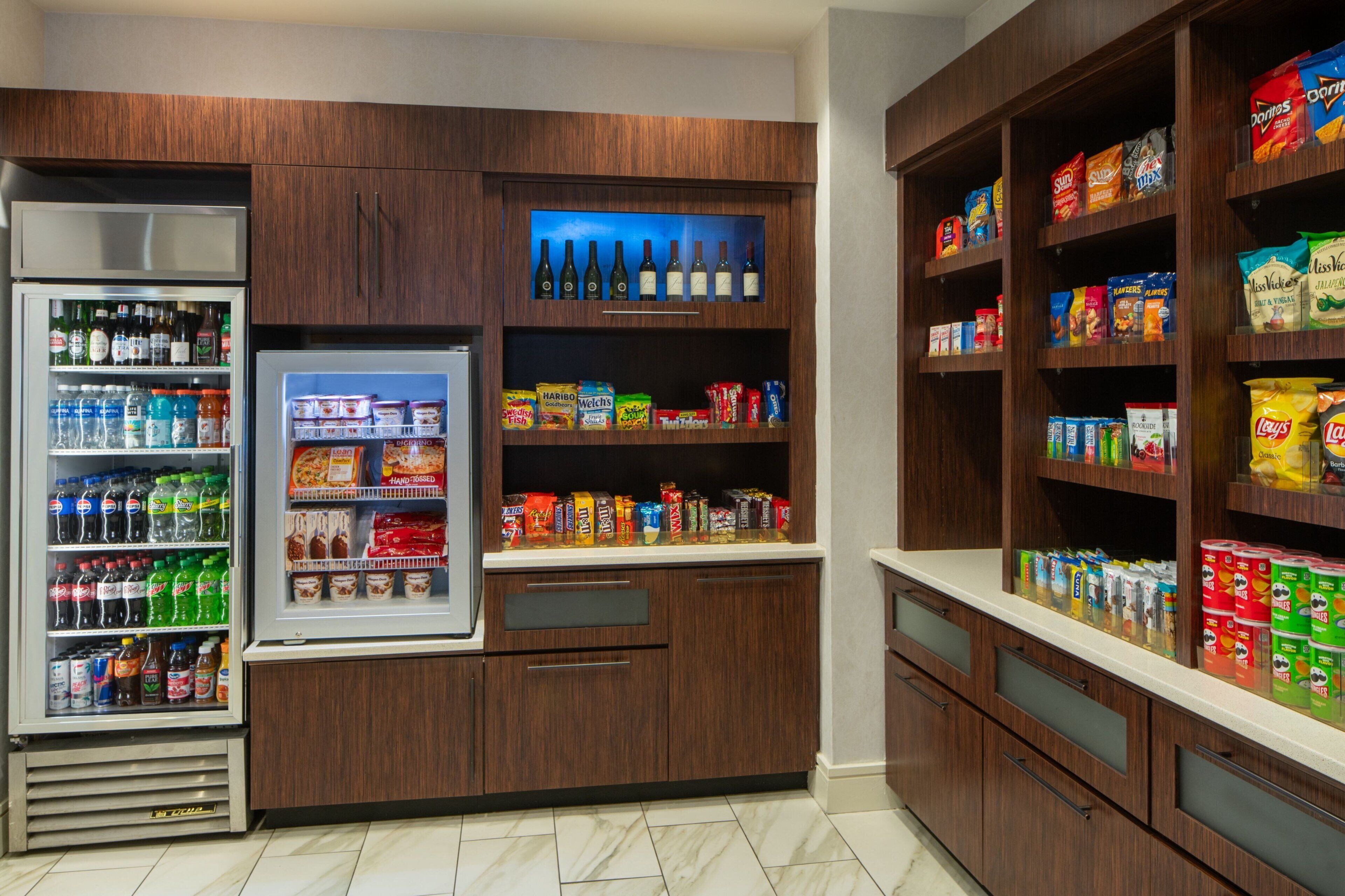 A kitchen with a refrigerator, shelves stocked with snacks, and a wine cooler.