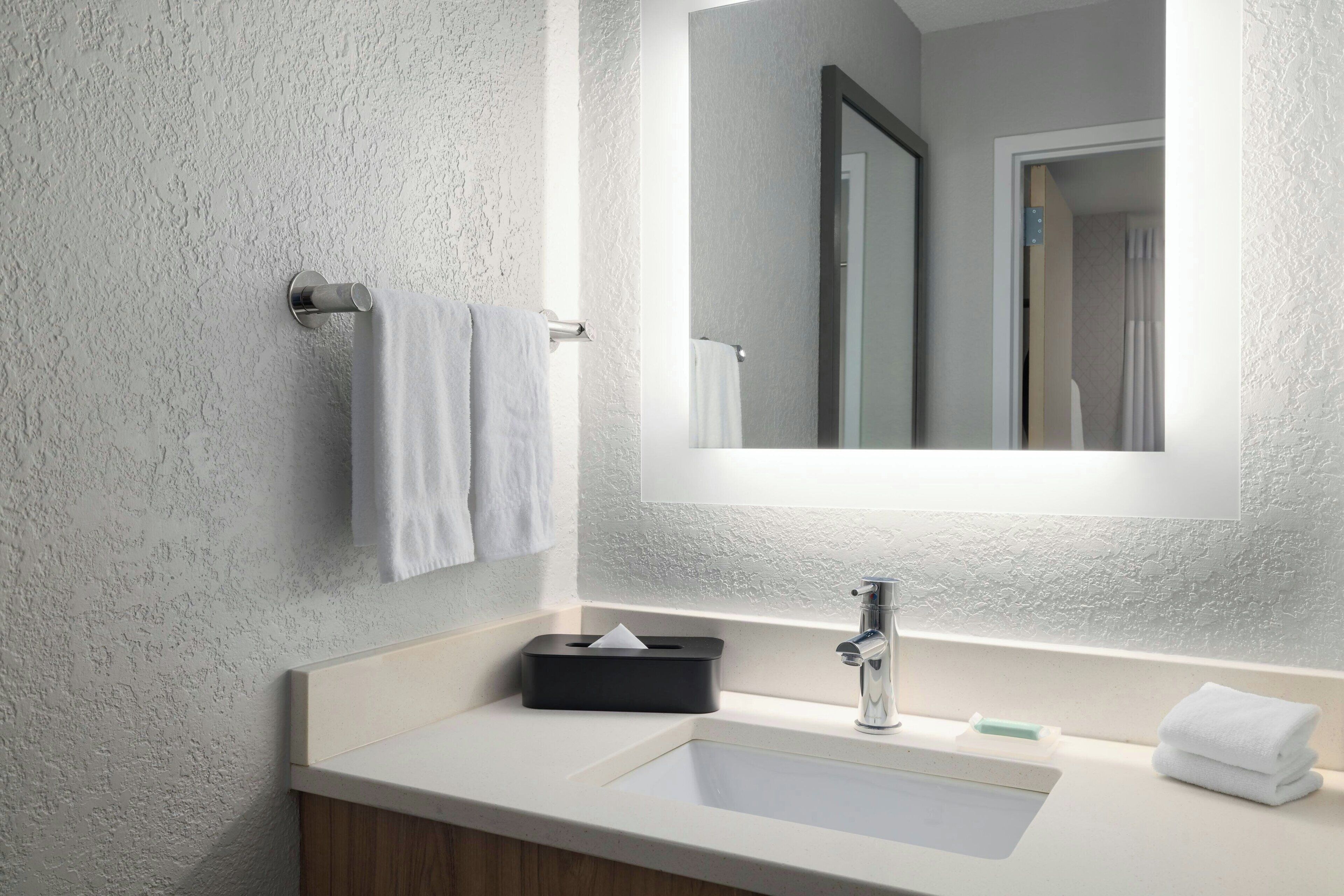 Une salle de bain moderne avec un mur blanc à texture, un grand miroir, un lavabo avec un robinet et deux serviettes suspendues à un porte-serviettes.
