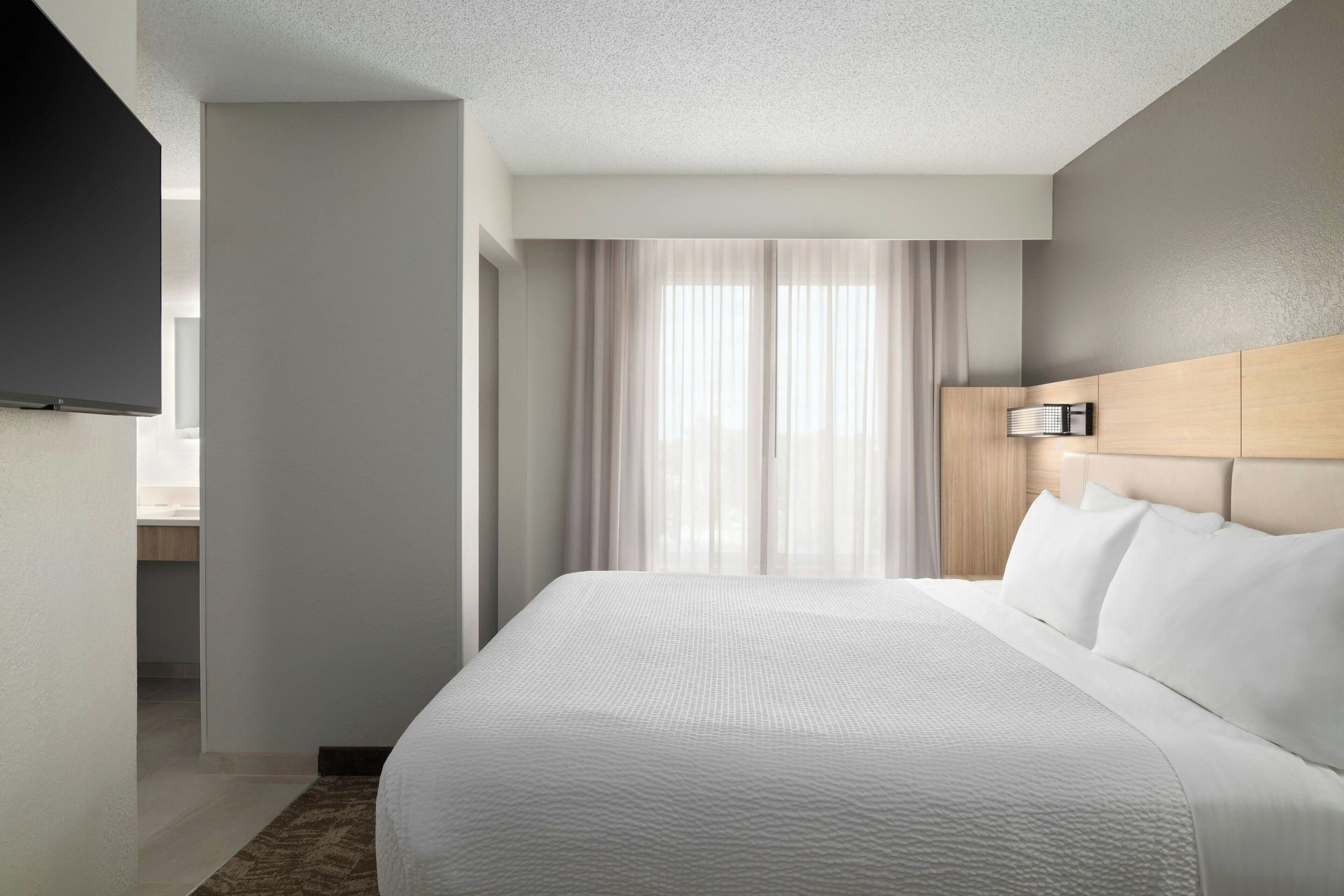 A modern hotel room with a large bed, a flat-screen TV mounted on the wall, and a large window with sheer curtains.