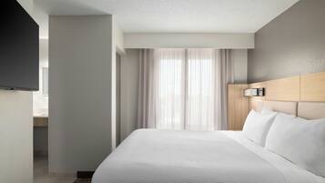 A modern hotel room with a large bed, a flat-screen TV mounted on the wall, and a large window with sheer curtains.