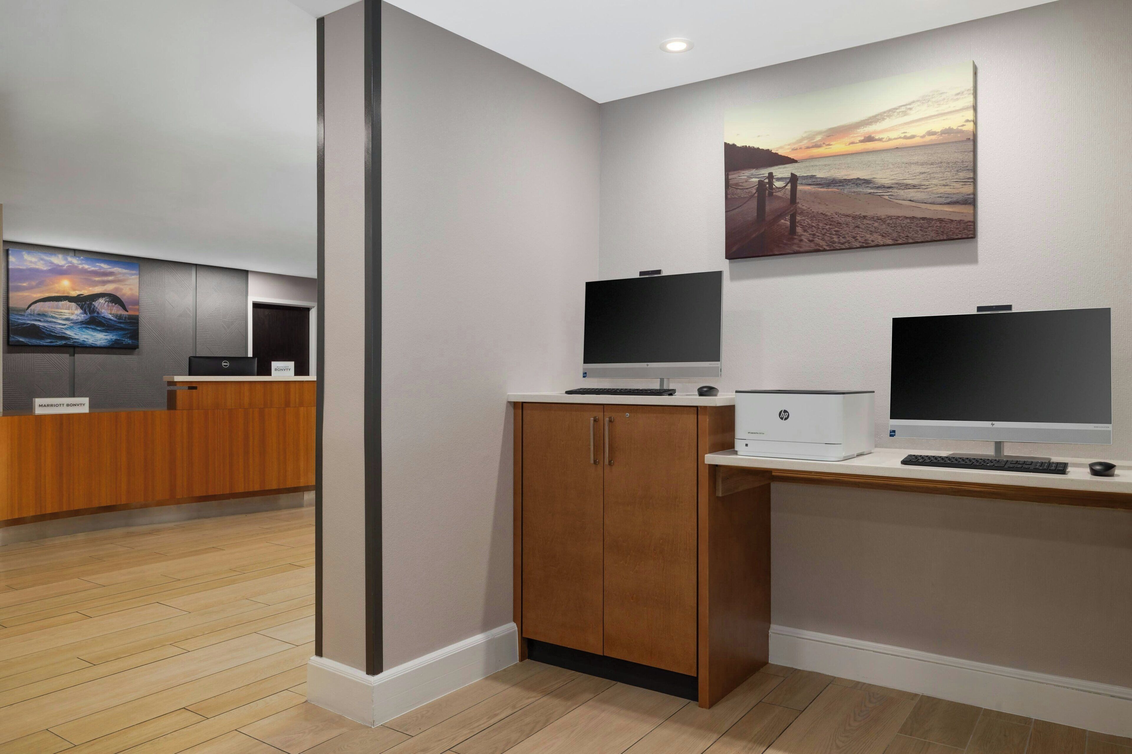 A modern hotel reception area with a wooden reception desk, two monitors, and a large painting of a sunset over the ocean.