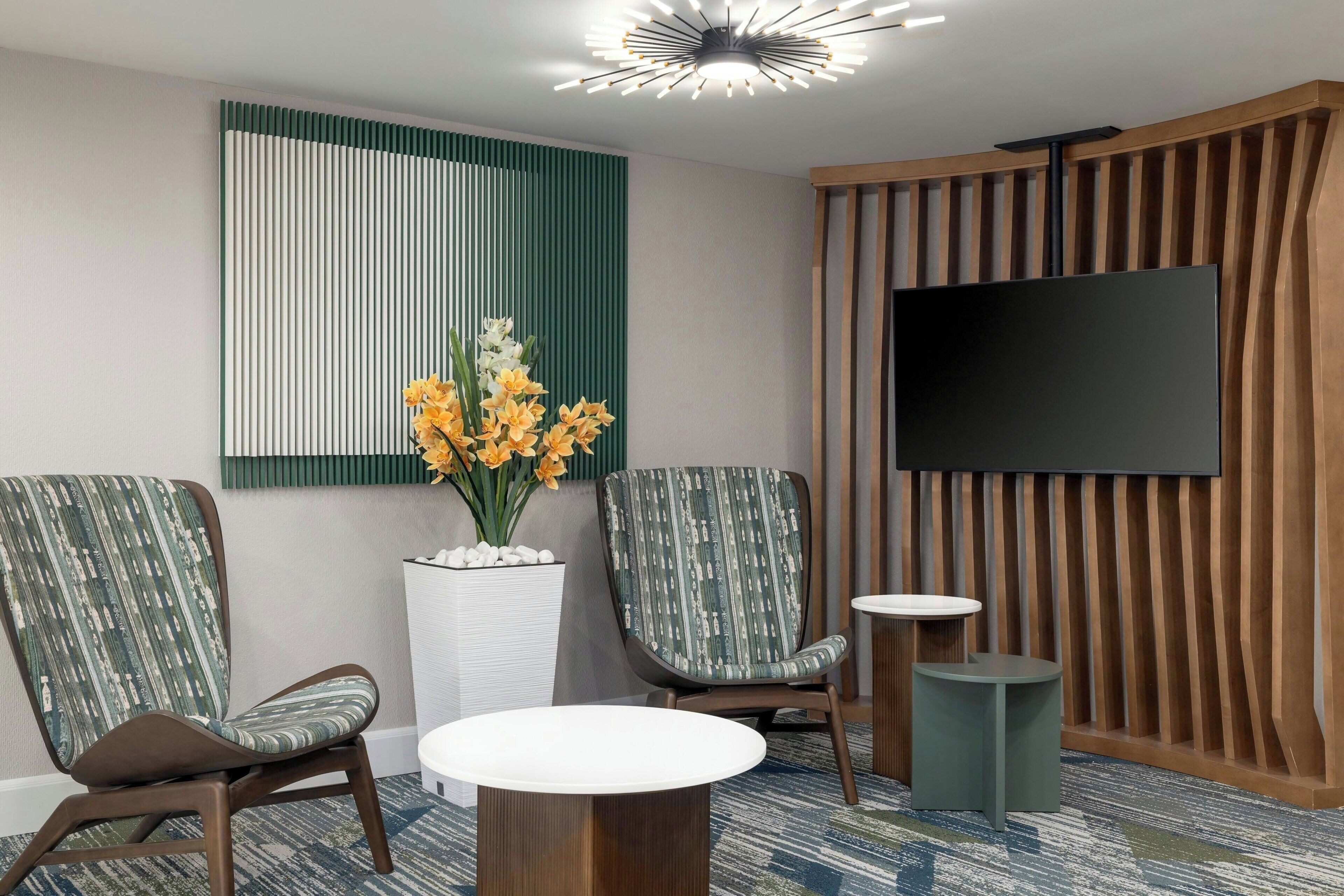 Un área de espera moderna con dos sillas estampadas, una mesa redonda, una planta en maceta con flores amarillas y un televisor de pantalla plana montado en una pared de paneles de madera.
