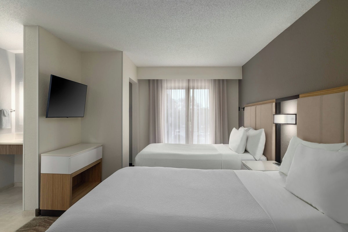 A hotel room with two beds, a TV mounted on the wall, a desk, and a window with curtains.
