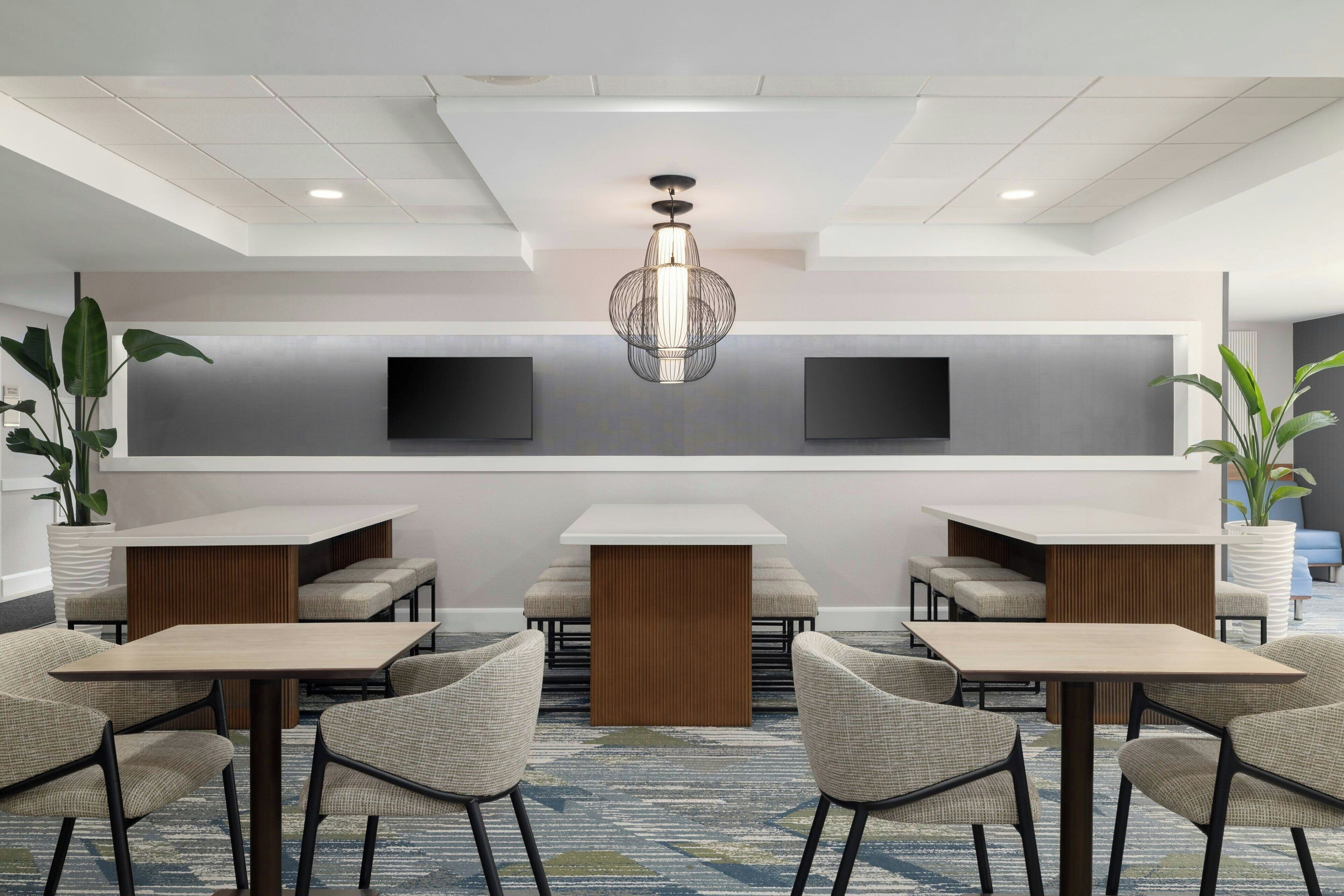 Um saguão moderno de hotel com área de estar central, um candelabro grande e duas TVs de tela plana na parede.