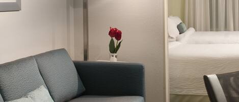 A hotel room with a grey sofa, a bedside table with a lamp, a vase with a red flower, and a bed with white bedding.